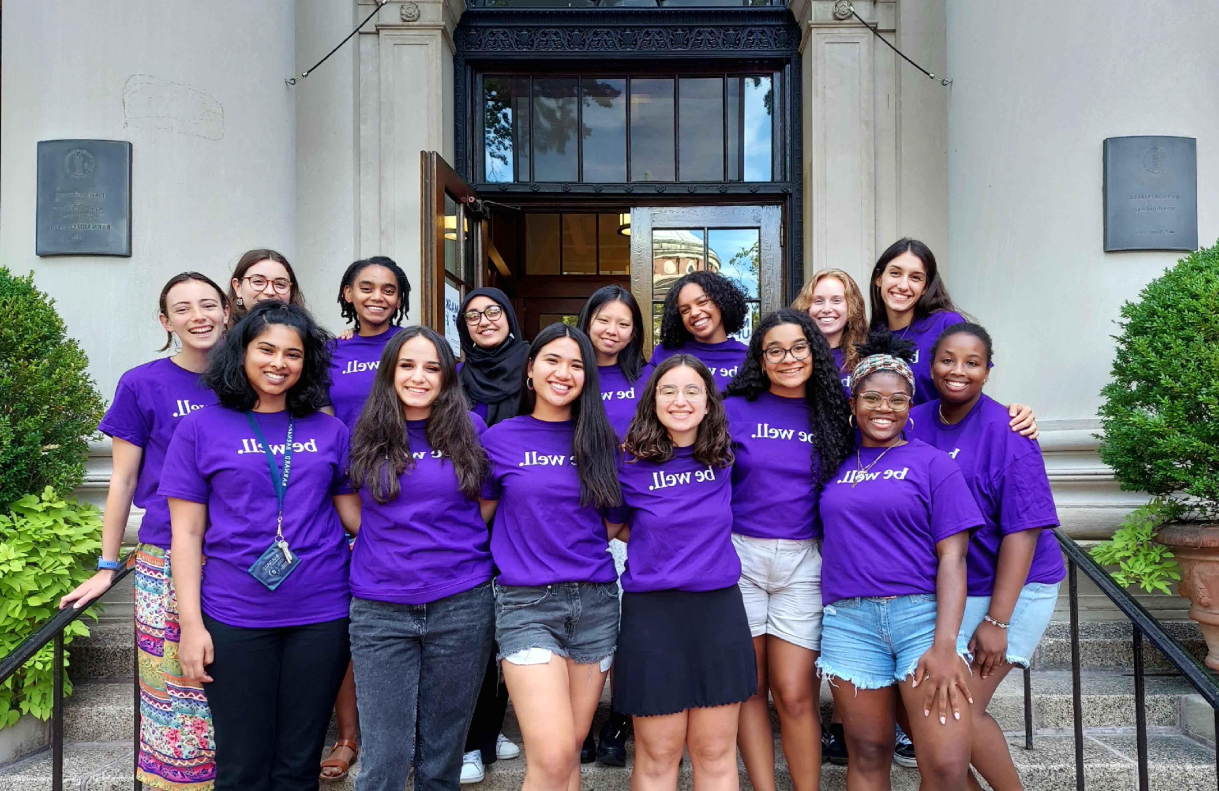 The Wellness Spot peer educators stand on the steps of Barnard Hall wearing purple t-shirts that say "be well" in white letters.